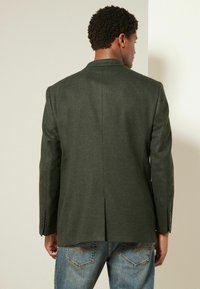 Olivgrüner Blazer, mit einem Schlitz auf der Rückseite, taillierte Passform, aus strukturiertem Stoff und mit Knopfärmel, kombiniert mit blauen Jeans.