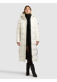 Ilgas baltas pūkinis paltas su kapišonu, juodos spalvos užtrauktuko detalėmis ir siūtu raštu. Dėvimas ant juodo viršutinio drabužio ir sijono.