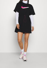 Svart kortärmad klänning med en rosa och vit Nike-logotyp, elastisk midja, och ihop med vita sneakers och strumpor.