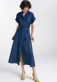 Jeansowa sukienka koszulowa z krótkimi rękawami, paskiem w talii i zapięciem na guziki z przodu. Posiada rozcięcie z przodu oraz swobodny design o długości midi.