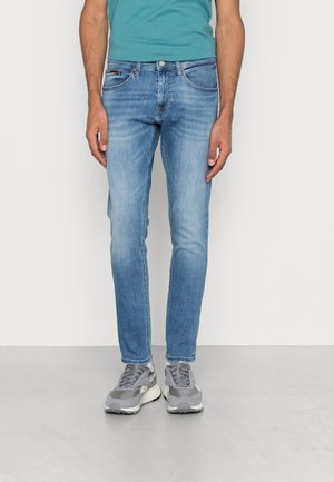 Mann trägt hellblaue Slim-Fit-Jeans, türkisfarbenes Hemd und graue Turnschuhe, steht vor einem schlichten weißen Hintergrund.