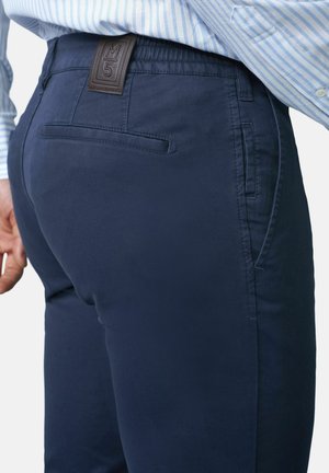 Man wearing navy blue pants and a light blue striped shirt, showing back pocket and branded leather patch on waistband.