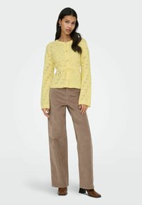 Cardigan jaune en maille avec motif ajouré, boutonné à l'avant, ceinturé à la taille, associé à un pantalon large en velours côtelé marron et des bottes marron à talons.