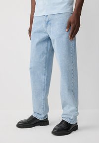 Jean en denim bleu clair avec une coupe décontractée, présentant une jambe droite, une texture décolorée et des poches avant. Associé à des mocassins noirs.