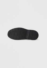 Suela de zapato de goma negra con ranuras horizontales, que presenta una forma plana y una textura suave. La marca está sutilmente grabada en el costado.