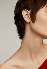 Pendiente de aro dorado con acentos verdes, diseño pequeño y delgado. El cabello corto del modelo enmarca el pendiente, con un fondo neutro.