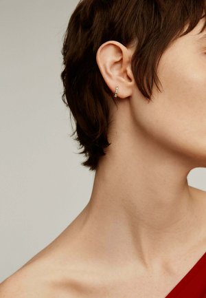 Pendiente de aro dorado con acentos verdes, diseño pequeño y delgado. El cabello corto del modelo enmarca el pendiente, con un fondo neutro.