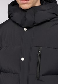 Svart vadderad jacka med hög krage, silverfärgade tryckknappar och en dragkedjeficka med läderdetalj. Slät, quiltad struktur synlig.