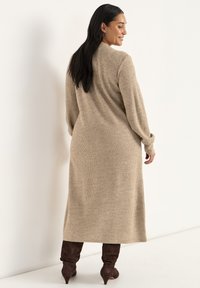 Beige neulemekko, jossa on pitkät hihnat, ribbitettu pinta ja nilkkapituus. Yhdistetty ruskeisiin polvipitkiin saappaisiin. Yksinkertainen ja vartalonmyötäinen muotoilu.