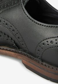Zwarte leren schoen met brogue-details, gestructureerd oppervlak, gebogen ontwerp en een contrasterende bruine houten zool.