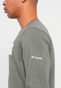 Mann i grå langermet skjorte som viser Columbia-logoen på overarmen, mot en nøytral lys bakgrunn, sett fra siden.