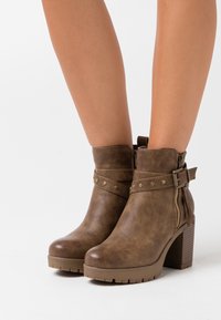 Stivali ankle in pelle marrone con tacco largo, cerniere laterali e cinturini decorativi con borchie in metallo indossati su gambe nude.