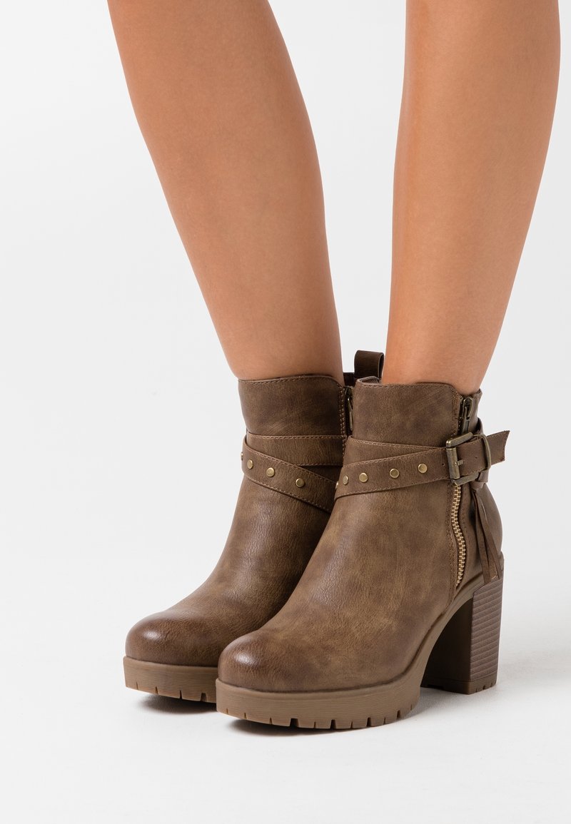 Stivali ankle in pelle marrone con tacco largo, cerniere laterali e cinturini decorativi con borchie in metallo indossati su gambe nude.