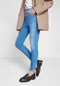 Persona vestida con jeans skinny de color azul claro, blazer beige, camisa blanca y mocasines negros, de pie con una mano en el bolsillo contra un fondo blanco.