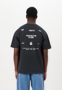 Black cotton t-shirt with crew neck, featuring printed text in white on the back: "OBEY," "AROUND THE GLOBE," and various city names.
