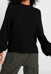 Pull noir en maille avec une texture côtelée, col rond et manches ballon. Porté avec un pantalon cargo gris, affichant un style ample.