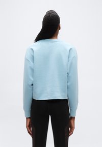 Γυναίκα με μακριά μαλλιά που φοράει ανοιχτό μπλε φούτερ και μαύρα κολάν, εμφανιζόμενη από πίσω μπροστά σε απλό ανοιχτό φόντο.