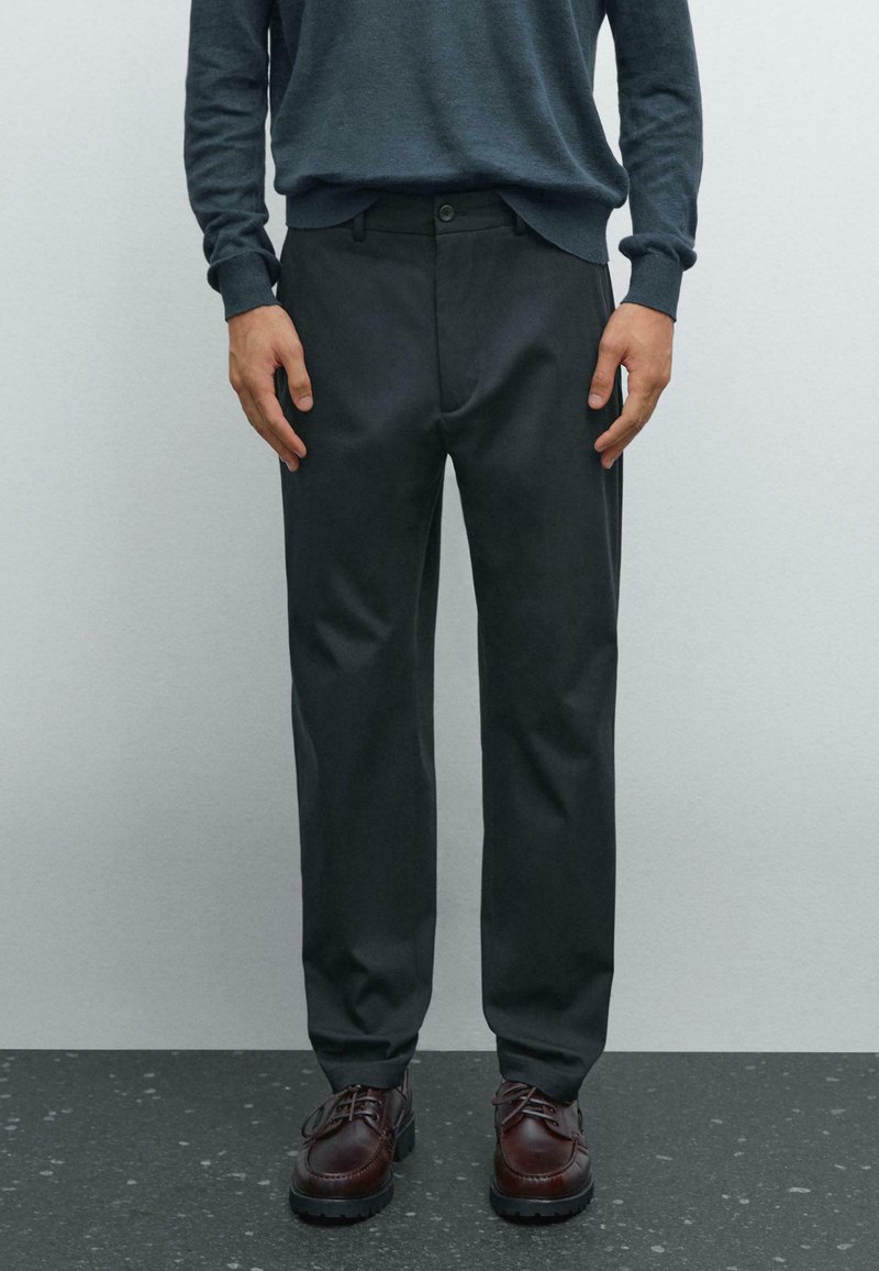 Pantaloni grigio scuro in tessuto liscio, con passanti per cintura, chiusura a bottone e tasche laterali, abbinati a scarpe stringate marroni.