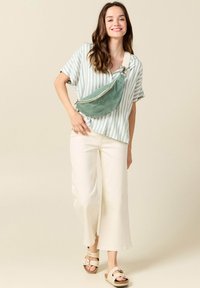 Borsa a tracolla di tessuto verde con cerniera, indossata a tracolla. Modello in una camicia a righe a maniche corte e pantaloni bianchi svasati, abbinata a sandali color sabbia.