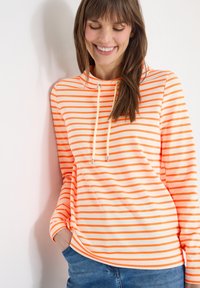 Femme souriante aux longs cheveux bruns portant un sweat à capuche orange et blanc rayé, appuyée contre un mur blanc avec une main dans la poche de son jean bleu.