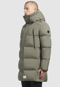 Jachetă puffer de culoare verde măslin, cu un guler înalt, buzunar lateral cu fermoar și patch cu branding pe tiv. Material neted, izolat.