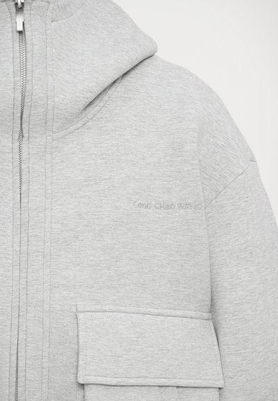 Sweatshirt gris clair avec capuche, comportant une fermeture éclair à l'avant, une grande poche kangourou et un logo subtilement en relief sur le tissu.