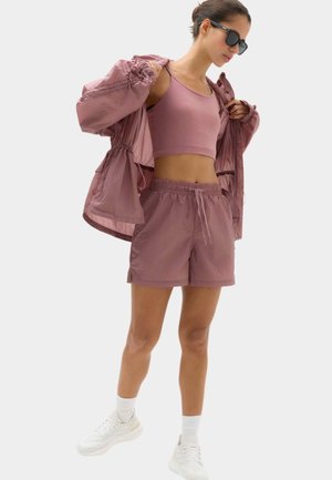 Kvinde iført matchende lilla sports-bh, shorts, let jakke, hvide sokker, sneakers og sorte solbriller mod en ensfarvet baggrund.