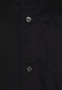 Tissu de chemise noire avec deux boutons gris à quatre trous et un bord de poche cousu visible sur le côté droit.