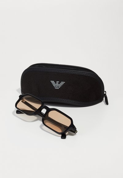Occhiali da sole neri con montatura ottagonale e lenti marroni, accompagnati da un astuccio nero con zip che presenta un logo sul fronte.