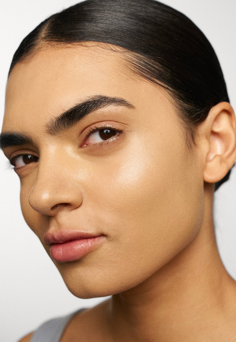 Portrait en gros plan d'une personne avec un teint lisse et brun clair, un maquillage subtil, des sourcils bien dessinés et des cheveux noirs et brillants plaqués en arrière.