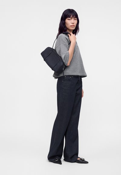 Femme portant un haut gris à manches courtes, un pantalon large noir, des chaussures noires pointues et portant un sac à épaule noir sur fond blanc.