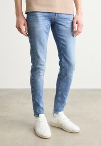 Jeans ajustados de color azul claro en tela de denim con áreas descoloridas. Combinados con zapatillas blancas que presentan un logo distintivo y un diseño minimalista.