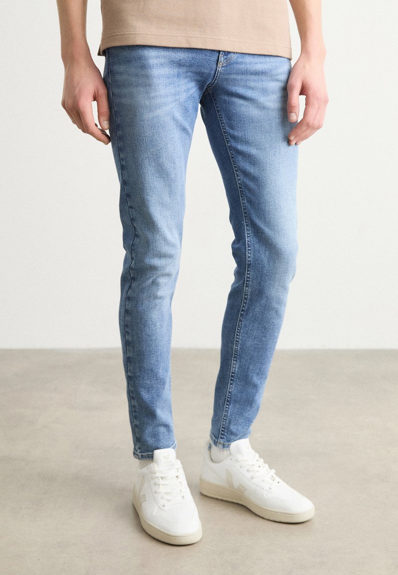 Jeans ajustados de color azul claro en tela de denim con áreas descoloridas. Combinados con zapatillas blancas que presentan un logo distintivo y un diseño minimalista.