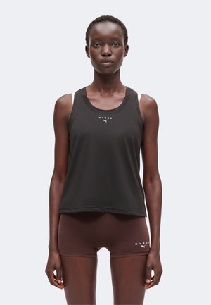 Ung kvinde stående med fronten mod kamera iført sort ærmeløs top og brune shorts med 'HYROX' og Puma-logoer på hver.