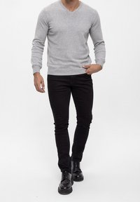 Homme portant un pull à manches longues gris clair, un pantalon noir slim et des chaussures en cuir noires à lacets, debout avec une main dans la poche.
