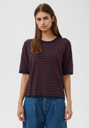 Jeune femme aux longs cheveux bruns portant un haut bleu marine à manches courtes avec des rayures rouges et un jean bleu ample, debout devant un fond uni.