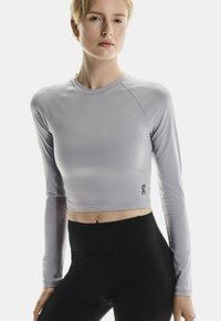 Hellblauer Langarm-Crop-Top aus dehnbarem Material mit rundem Halsausschnitt und schwarzem Logo-Akzent auf der Vorderseite. Kombiniert mit schwarzen Leggings.