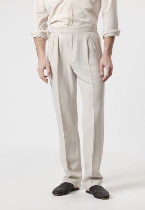 Hombre que lleva pantalones plisados de color crema, camisa de manga larga a juego con las mangas remangadas, y zapatos negros tejidos sin cordones.