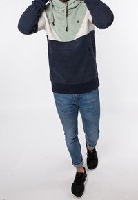 Felpa con cappuccio in blu navy, grigio chiaro e verde menta, con tasca frontale e cordino regolabile. Indossata con jeans blu e sneaker nere.