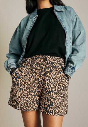 Personne portant une veste en jean bleue, un haut noir et un short imprimé léopard, les mains dans les poches devant un fond uni.