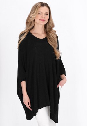 Blond žena v loose černém ponču s tříčtvrtečními rukávy a bílými kalhotami, stojící před bílým pozadím.