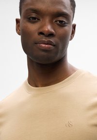 Camiseta de cuello redondo color beige hecha de una tela suave. Presenta un sutil logo bordado en el lado izquierdo del pecho y un cuello sin costuras.