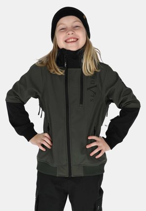 Smilende barn iført sort hue, olivengrøn lynlåstrøje med sorte ærmer og sorte bukser, stående med hænderne på hofterne.