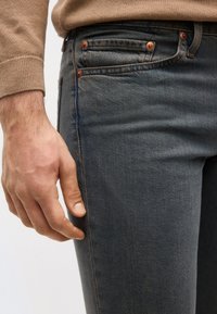 Κοντινό πλάνο του χεριού ενός ατόμου που ακουμπάει δίπλα σε σκούρο μπλε τζιν, σε συνδυασμό με μανίκι μπεζ πουλόβερ.