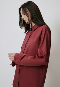 Camicia rossa con bottoni, dal tessuto liscio, maniche lunghe e colletto. Presenta dei sottili dettagli delle cuciture e polsini regolabili.