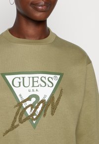 Sudadera verde oliva con un logo triangular blanco que presenta "GUESS U.S.A." y texto "Icon" adornado en acentos dorados. Textura de tela suave.