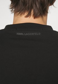 T-shirt nera con texture a coste. Presenta un collo rotondo e un logo goffrato sottile "KARL LAGERFELD" nella parte posteriore vicino al colletto.
