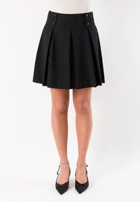 Gonna nera plissettata con una larga fascia in vita, caratterizzata da due bottoni e una texture liscia, lunghezza sopra il ginocchio. Abbinata a décolleté nere con punta a punta.