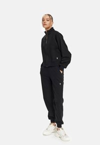 Schwarze kurz geschnittene Zip-Jacke mit hohem Kragen und elastischen Bündchen, kombiniert mit passenden Jogginghosen mit Seiten Taschen und dezenten Logoakzenten.