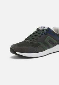 Men's athletic shoe in dark gray and green, featuring a textured mesh upper, reinforced toe, and white sole with subtle accents.